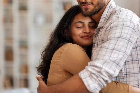 Happy Indian couple in love hugging, cuddling at home, woman closing eyes and smiling, enjoying tender moment with husbandの写真素材