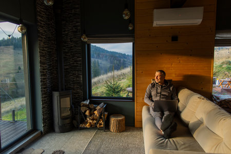 Man sitting on couch in house, using laptop and looking at window with scenic view of mountains,...
