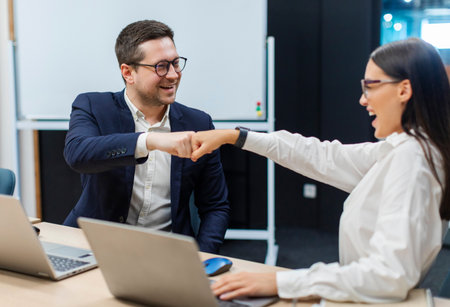 Joyful two colleagues bumping fists, greeting each other or showing team union, business partners celebrating shared success, sitting in officeの写真素材