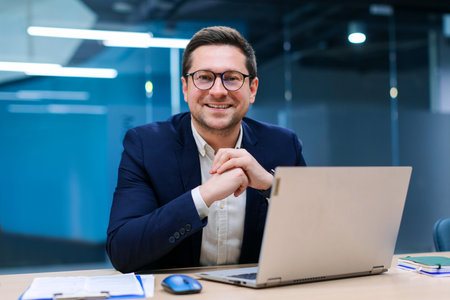 Charismatic European businessman sitting at desk in stylish office working on laptop and ready for...