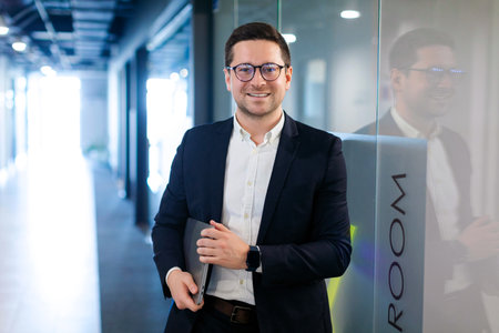 Business person. Portrait of European businessman with laptop in hands standing in office hallway,...
