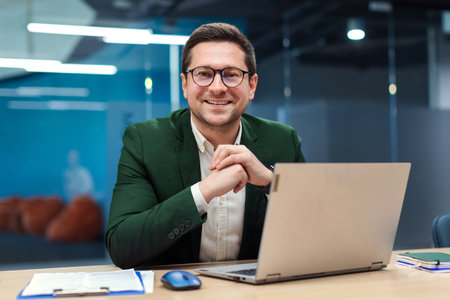 Charismatic European businessman sitting at desk in stylish office working on laptop and ready for...