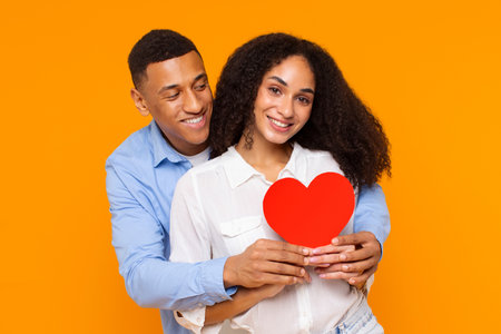 Valentines Day. Portrait of happy black couple holding red paper heart and hugging, posing on yellow...
