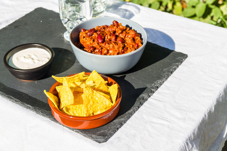 Home made Chilli Con Carne in orange ceramic bowl and tortilla chips and soured cream on the side with jug and glass of iced water. On slate mat, outside on linen covered table. Evening lightの写真素材