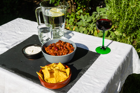 Home made Chilli Con Carne in orange ceramic bowl and tortilla chips and soured cream on the side with jug and glass of iced water. On slate mat, outside on linen covered table.の写真素材