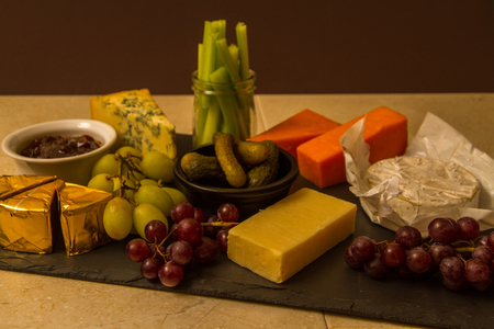 Cheeseboard, platter for sharing, cheese, red Leicester, stilton, cheddar, camembert, brie, grapes, pickled gherkins, celery, pickle or chutney on slate.の写真素材