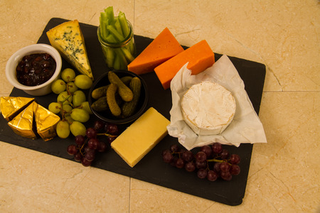 Cheeseboard, platter for sharing, cheese, red Leicester, stilton, cheddar, camembert, brie, grapes, pickled gherkins, celery, pickle or chutney on slate.の写真素材