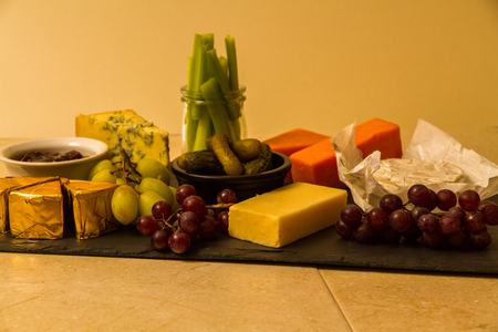 Cheeseboard, platter for sharing, cheese, red Leicester, stilton, cheddar, camembert, brie, grapes, pickled gherkins, celery, pickle or chutney on slate.の写真素材