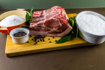 Pork loin or back joint with ingredients for dry curing into bacon, salt, sugar, peppercorns, juniper berries and bay leaves, landscape, on wooden chopping board, copyspace at bottom.の写真素材