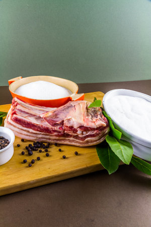 Pork belly  joint with ingredients for dry curing into bacon, salt, sugar, peppercorns, juniper berries and bay leaves, portrait.の写真素材