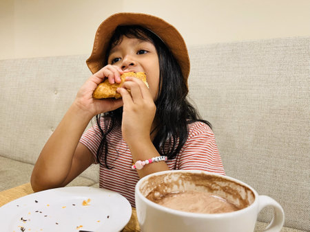 Asian little girl eating a chicken pastel and drinking delicious hot cocoa or chocolate drinking in white cup and sloppy for breakfast in the morning on table cafe restaurantの写真素材
