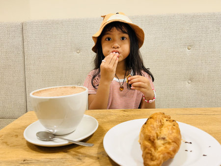 Asian little girl eating a pastry with fully mouth and drinking delicious hot cocoa or chocolate drinking in white cup and chicken pastel for breakfast in the morning on table cafeの写真素材