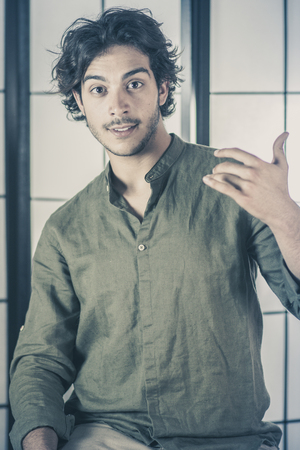 An handsome young man, 20Y, is posing in studio with raised hands at chest with interrogative expression. Green skirt. Retroilluminated booths in the background.の写真素材