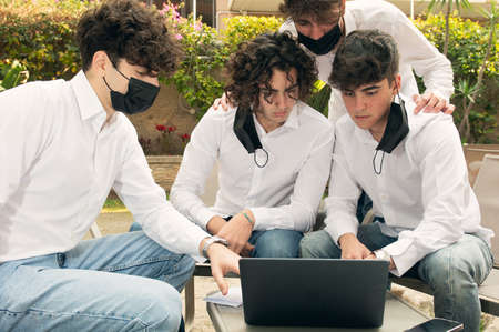 A group of young students are sitting at a table, working at a laptop, they are wearing masks for Covid prevention but some have face uncovered .の写真素材