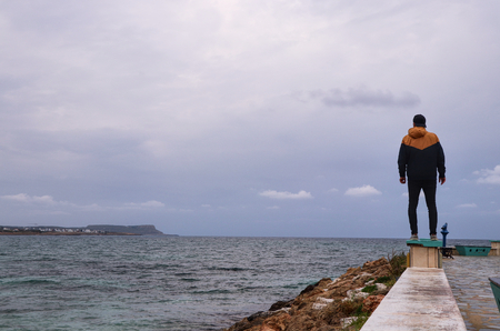Young man standing on the edge wharf and watching mountains on other side. Man is standing on the edge of Ayia napa city which is formed from a smaller island just near the main shoreの写真素材