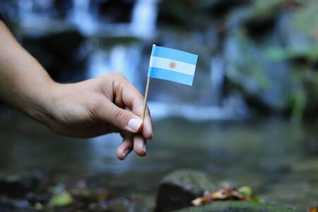 Argentina in the hands of powerful people. Young man holds flag of Argentina near stream. Concept of humanity and dominance. Prove of depraved and avariciousness. Colour man waves with flag.の写真素材
