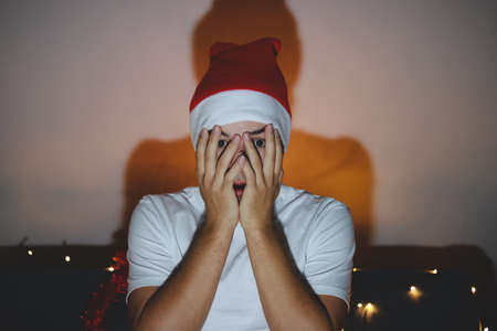 young man covers his face with his hands out of fear. Gentleman sitting on couch and watching horror movie. Fear, feeling hopeless. Christmas moment of surprise and horror.の写真素材