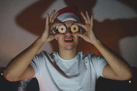 Funny expression of a young man with a Christmas hat. Man holding Christmas cookies instead of eyes....