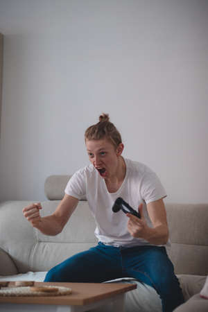 young man in a white T-shirt and a pigtail on his head rejoices in the game console won in his...