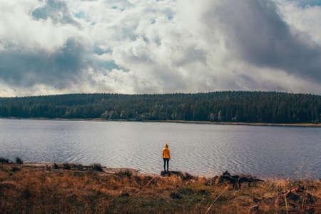 young girl in yellow jacket stands on shores of Lake Bedrichov in Jizera Mountains. Concept of wild nature and innocent girls. Candid portrait of woman in distinctive yellow jacket in antique whiteの写真素材