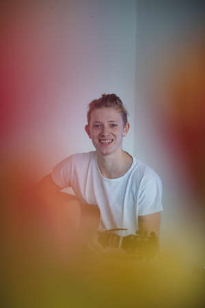 real smile of a young singer in a white T-shirt sitting in the middle of the room. the smile of playing an acoustic guitar. The artist smiles while doing his profession on his afternoon off.の写真素材
