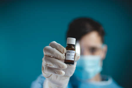 doctor in a protective shield, veil, and sterile gloved suit holds a single weapon against the Coronavirus pandemic. A scientist holds an ampoule against a Covid-19 and points to the camera.の写真素材