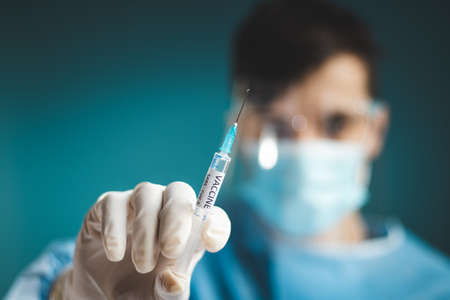 doctor in a sterile protective suit with a shield on his head prepares a syringe with a Covid-19 vaccine for an incoming patient. Medical worker. Fighting a pandemic. Blue tone.の写真素材