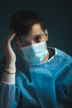 Torn young doctor after work in intensive care at the hospital. A twelve-hour shift full of acute cases collects a tax. A doctor dressed in sterile clothing and a shield against Covid-19 infection.の写真素材
