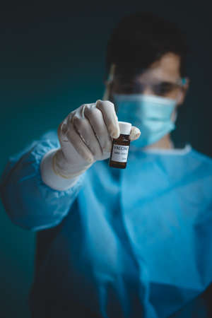 doctor in a protective shield, veil, and sterile gloved suit holds a single weapon against the Coronavirus pandemic. A scientist holds an ampoule against a Covid-19 and points to the camera.の写真素材