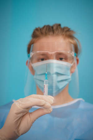 doctor in a sterile protective suit with a shield on his head prepares a syringe with a Covid-19 vaccine for an incoming patient. Medical worker. Fighting a pandemic. Blue tone.の写真素材