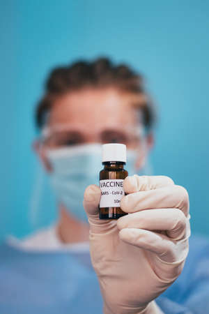 doctor in a protective shield, veil, and sterile gloved suit holds a single weapon against the Coronavirus pandemic. A scientist holds an ampoule against a Covid-19 and points to the camera.の写真素材