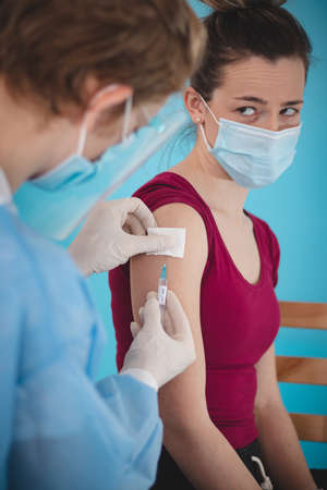 doctor in sterile and protective clothing injects a covid-19 vaccine into the shoulder of a young woman with a worried expression. Vaccination time. Global pandemic.の写真素材