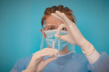 doctor in a sterile protective suit with a shield on his head prepares a syringe with a  vaccine for...