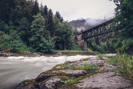 Ancient green railway bridge over wild river Enns in rainy weather in Gesause National Park near town of Admont in center Austria. Long exposure emanates milk from water flowing along green nature.の写真素材
