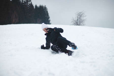 young boy with a white cap laughs at his own stupidity. The teenager was riding on a ski slope on a plastic board and landed hard. Laugh at your own mistakes. Snow fooling around in childhood.の写真素材