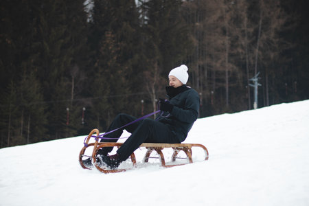 Tobogganing in winter at dizzying speed on the piste with a smile on your face. Kids' joy. A boy in a black jacket and white hat rides on a wooden sledge from times past. Enjoying winter.の写真素材