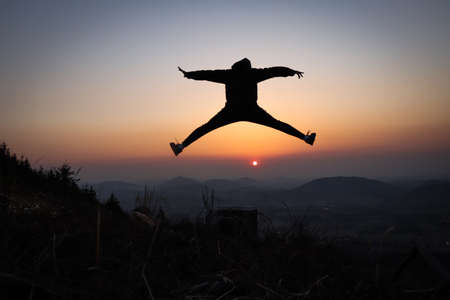 Silhouette of a teenager and athlete jumping on a tree stump and doing a straddle vault, splits at sunset in the European mountains. Training in natural conditions. Self weight training. Coordination.の写真素材