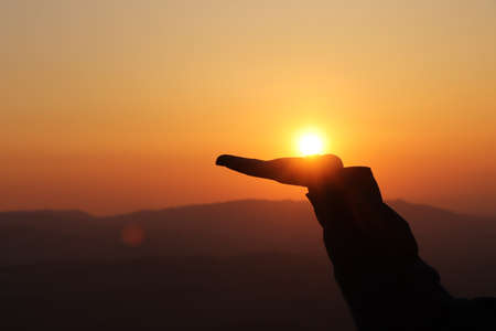 Hand from a man tries to hold on to the palm of the setting sun on top of a mountain in the Beskydy Mountains, Czech Republic. Catching the sun in your hand. Hot orange ball on the palm.の写真素材