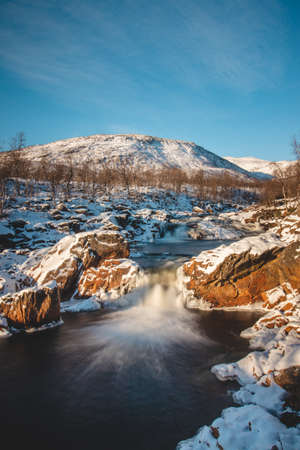 Winter fairy tale in the form of Storforsen waterfall in Silsand, Senja, Norway. Sunset illuminates the wild waterfall. In the background, the snowy hills of the Norwegian landscape.の写真素材