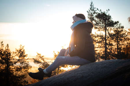 brunette woman in blue jeans and a black coat sits on top of a rock at sunrise looking down into the...
