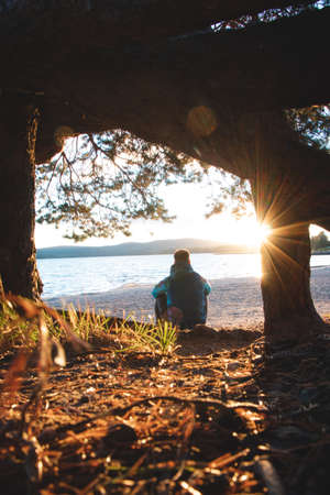 Pensive hiker sits on a sandy beach after climbing a mountain, looking out into the lake and...