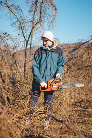 young 17-year-old temporary worker in work clothes wrestles with a chainsaw and a dry larch tree....