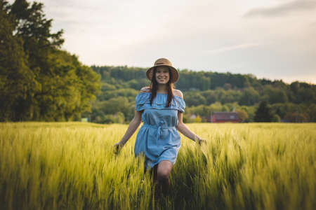 Breathtaking candid portrait of a brunette aged 20-24 walks in a beautiful blue dress and hat in a cornfield, smiling naturally. Fashion vintage style. Natural beauty of a brown haired European woman.の写真素材