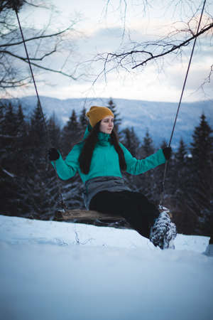 young lady swings on a swing in the winter months and enjoys the fresh air. Winter clothes and beautiful brunette. Act like a child.の写真素材