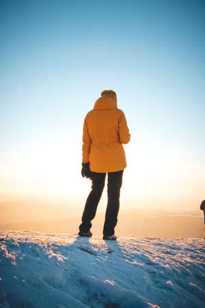 Traveler in a distinctive yellow jacket stands on top of a hill in the Polish Beskydy Mountains at sunset, enjoying the moment. Real people in winter ice environment.の写真素材