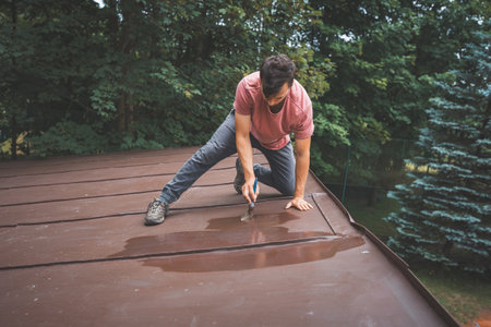 Man crouches down, carefully painting the surface of a roof with a small brush. His focus is on...