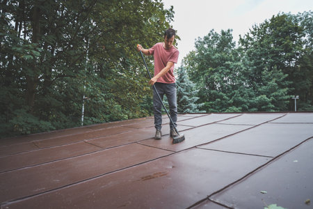 Man is cleaning a slanted roof with a long broom, surrounded by trees. The outdoor setting features lush greenery, with the individual focused on his task of removing debris from the roof.の写真素材