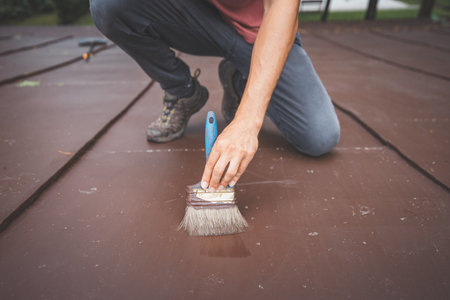 Close-up of a man crouching while painting a roof. His hand holds a brush, carefully applying a fresh coat of paint to the surface. The focus is on the detailed craftsmanship of the task at hand.の写真素材