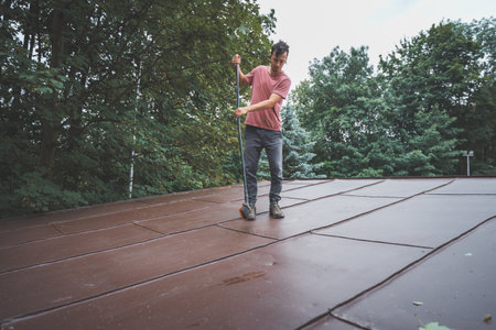 Man is cleaning a slanted roof with a long broom, surrounded by trees. The outdoor setting features lush greenery, with the individual focused on his task of removing debris from the roof.の写真素材