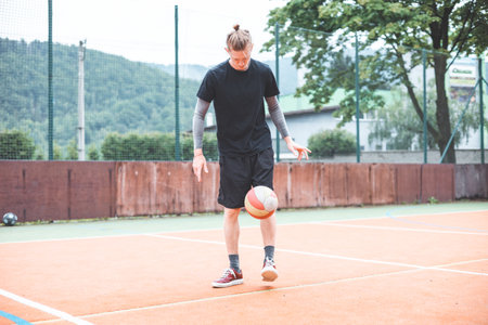 Young man casually juggles a football on an outdoor sports court, focusing on his footwork. Dressed...